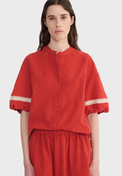 Jeune femme aux longs cheveux bruns portant un haut rouge boutonné avec des manches bouffantes et un détail en dentelle blanche, associé à un bas rouge assorti.