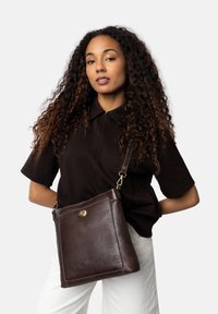 Borsa a spalla in pelle marrone con una superficie liscia, forma quadrata, dettagli in hardware dorato e tracolla regolabile.