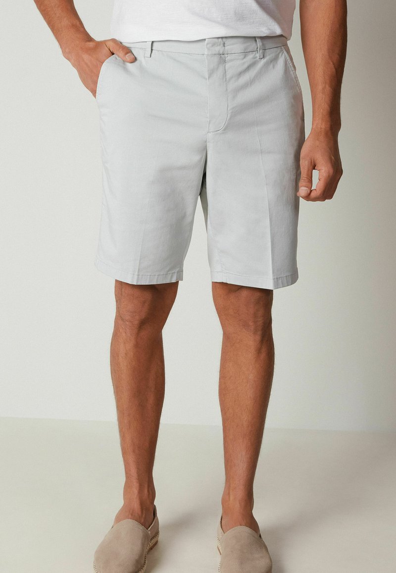 Shorts grigi chiaro realizzati in tessuto misto cotone, con tasche laterali, una linea dritta e una texture liscia. Indossati con scarpe beige senza lacci.