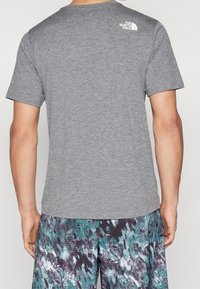 Šedé tričko s krátkými rukávy a jemnou texturou. Na zadní straně u výstřihu je malý bílý logo. Nositelný s vzorovanými plavkovými šortkami.