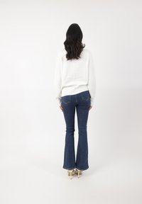 Suéter blanco con dobladillo acanalado, combinado con jeans acampanados de color azul oscuro que tienen bolsillos traseros. El modelo está de espaldas frente a un fondo blanco.