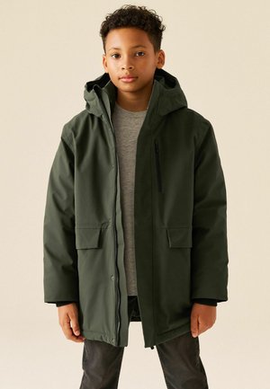 Ragazzo che indossa un cappotto invernale verde scuro con cappuccio sopra una maglietta grigia e pantaloni scuri, in piedi davanti a uno sfondo chiaro.