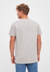 Man with short curly hair wearing plain light gray t-shirt and blue jeans, facing away against white background.