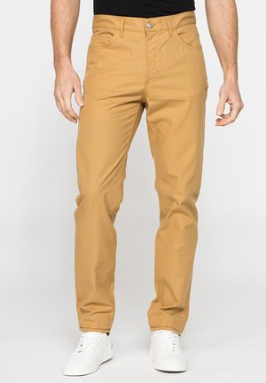 Uomo che indossa pantaloni beige slim fit con tasche frontali, passanti per cintura e sneakers bianche, in piedi davanti a uno sfondo semplice.