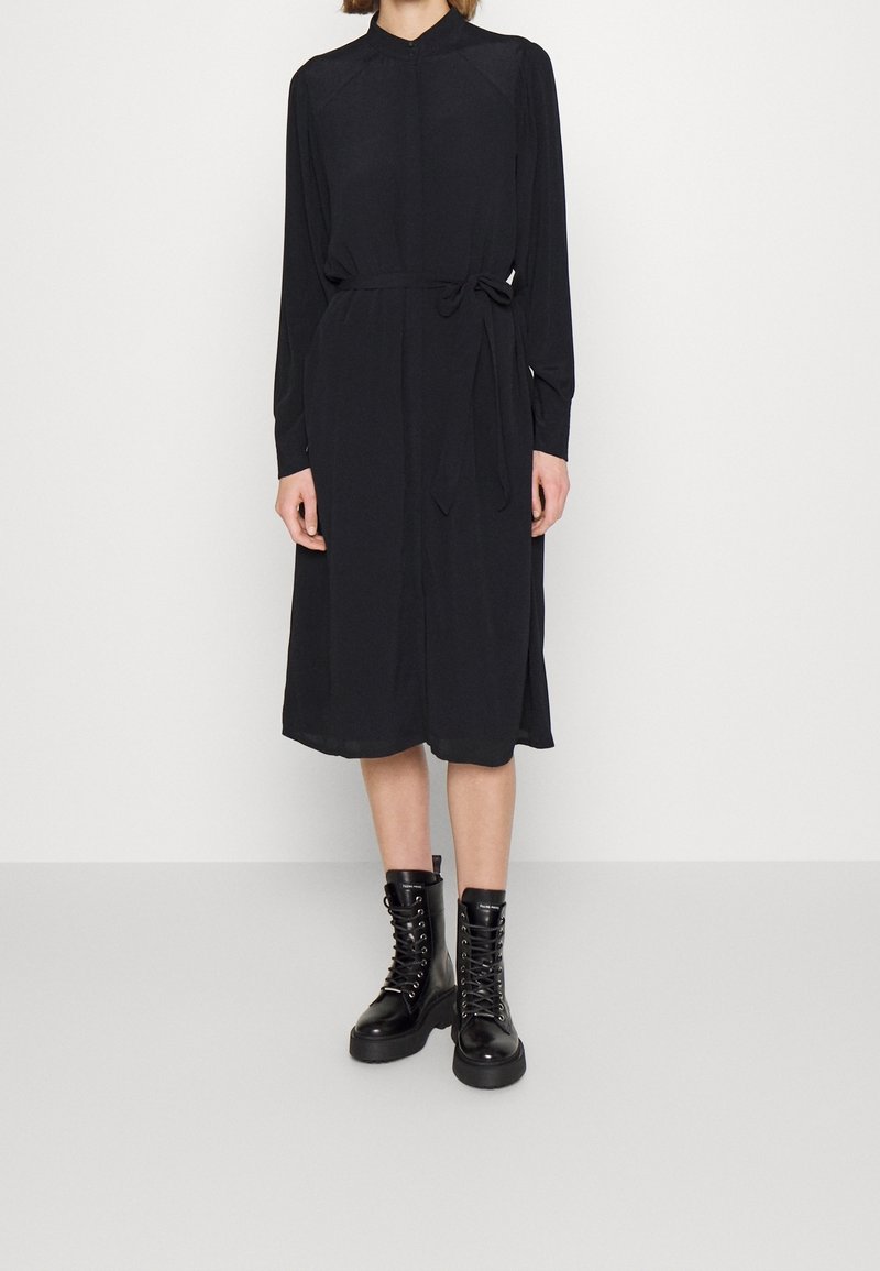 Personne portant une robe midi noire à manches longues avec un lien à la taille et des bottines noires à lacets, debout devant un fond blanc uni.