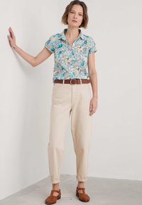 Cămașă cu guler, cu mânecă scurtă, având un model floral și geometric colorat, asortată cu pantaloni bej înguști și pantofi maro împletiți.