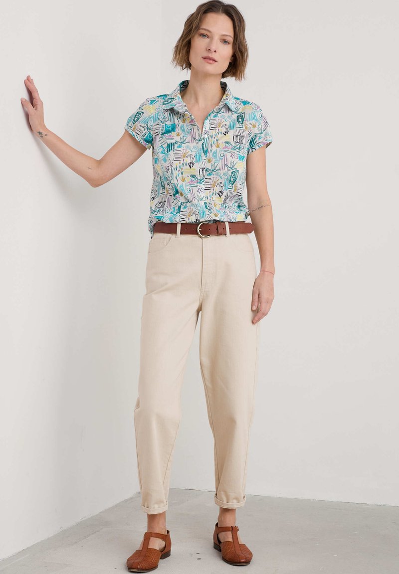 Cămașă cu guler, cu mânecă scurtă, având un model floral și geometric colorat, asortată cu pantaloni bej înguști și pantofi maro împletiți.