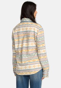 Flanellhemd mit einem gelben, cremefarbenen und blauen Muster aus geometrischen Formen und Streifen. Lange Ärmel und spitzer Kragen. Weiche Textur.