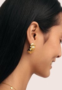 Boucles d'oreilles en or avec un design torsadé, présentant une surface lisse et réfléchissante et une esthétique contemporaine, portées sur l'oreille d'un modèle.