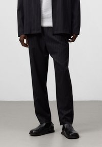 Uomo che indossa pantaloni neri a righe, scarpe slip-on nere, una giacca scura e un maglione bianco, in piedi su un pavimento grigio.