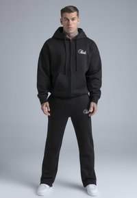 Zwarte capuchontrui met zakken aan de voorkant en wit logo, gecombineerd met bijpassende broek. Zachte, gestructureerde stof met een ontspannen pasvorm en trekkoorden.