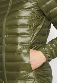 Dłoń osoby schowana w bocznej kieszeni błyszczącej, pikowanej oliwkowo-zielonej kurtki z zamek błyskawicznym z przodu i elastycznymi mankietami.