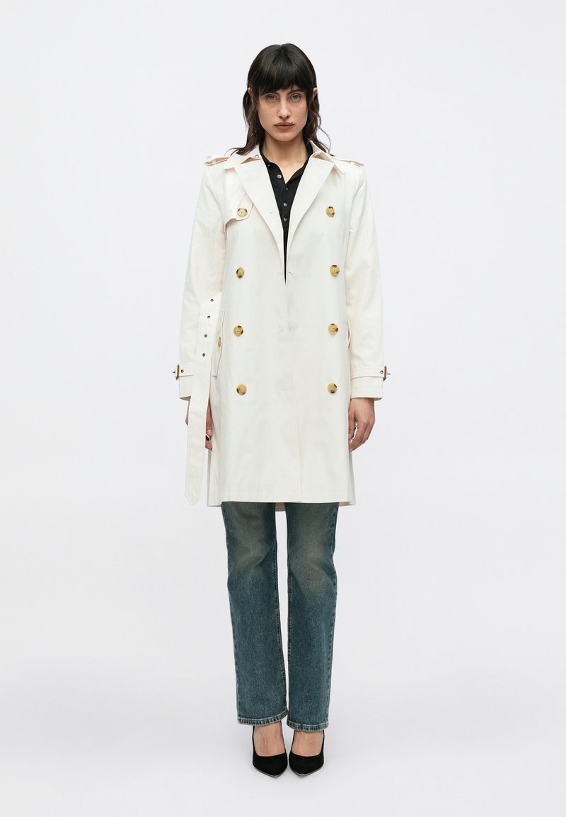 Fehér ballonkabát arany gombokkal, dupla soros kivitelben, vállpántokkal és megkötős derékkal. Fekete galléros póló és kék farmerruhával viselve.