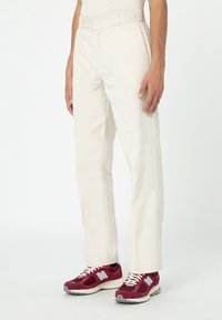 Mann in cremefarbenen, geraden Hosen und burgunderfarbenen New Balance Sneakers, der vor einem hellen, einfarbigen Hintergrund steht.