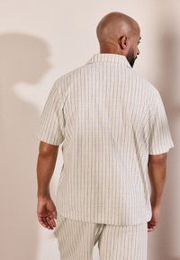 Camicia a maniche corte con colletto in beige chiaro con righe verticali. Il materiale è leggero e strutturato, abbinato a pantaloni coordinati.