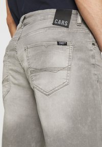 Ljust grå denimjeans med bakfickor, synlig sömnad och svarta märken med texten "CARS" på midjebandet och fickan.