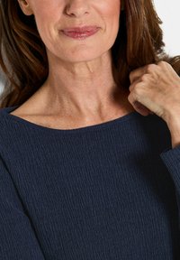 Navyblaues, geripptes Oberteil mit weitem Ausschnitt, aus strukturiertem Stoff und langen Ärmeln. Die Hände, die auf den Schultern ruhen, ergänzen das Detail.
