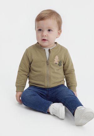 Lille barn sidder på gulvet iført en grøn jakke med lynlås og træpatches, blå jeans og hvide sneakers, kigger let til siden.