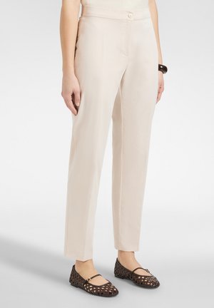 Frau trägt hoch taillierte beige Hose mit Knopfverschluss, kombiniert mit dunkelbraunen, gewebten Mary Jane Ballerinas und einem schwarzen Armband.