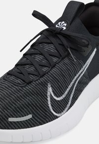 Černá sportovní bota s texturovaným svrškem, s bílým vyšitým logem swoosh, plochými tkaničkami a bílou gumovou podrážkou s horizontálními drážkami.