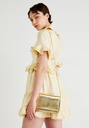 Joven con un vestido corto amarillo con volantes, llevando un pequeño bolso dorado con cadena sobre el hombro, mirando por encima de su hombro izquierdo.