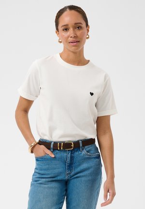 Femme à la peau brun clair et aux cheveux bouclés attachés porte un t-shirt blanc avec un petit cœur noir, un jean bleu, des boucles d'oreilles créoles dorées et un bracelet.