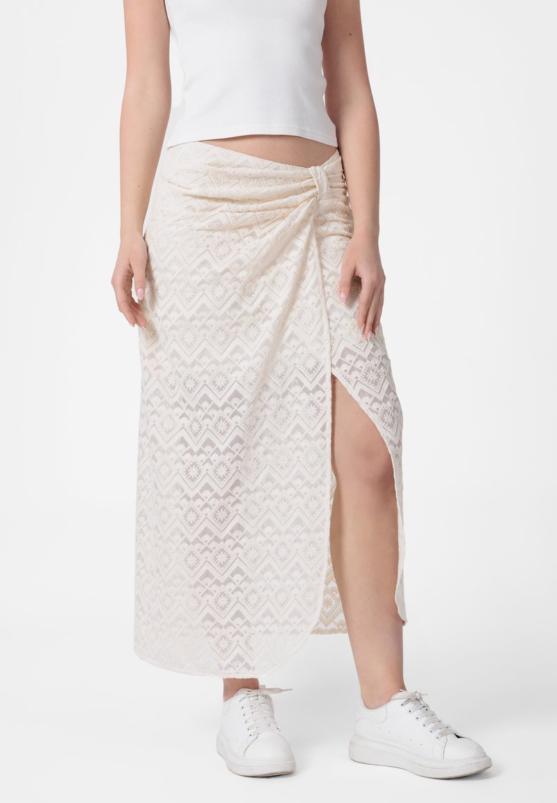 Maxi-jurk van kant in gebroken wit met een geometrisch patroon, knoopdetail bij de taille en een zijsplit. Draagt met witte sneakers.