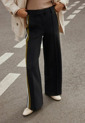 Person iført skræddersyede sorte vidde bukser med hvide og olivengrønne sidelommer, beige jakke og creme ballet flats stående på asfalt.