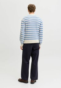 Personne debout, vue de dos, portant un pull rayé crème et bleu, un pantalon noir ample et des chaussures marron sur un fond uni.