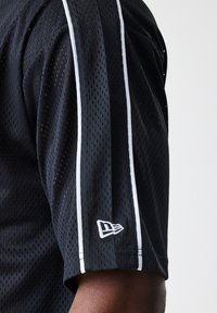 Svart mesh sportstrøye med hvite sidestiper og en logo på ermet. Stoffet er pustende med et perforert design.