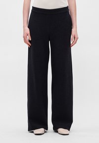 Platas kājas bikses tumšā audumā ar vertikālām svītrām, ar elastīgu jostu un gludu tekstūru. Saskaņotas ar bēšām kurpēm.