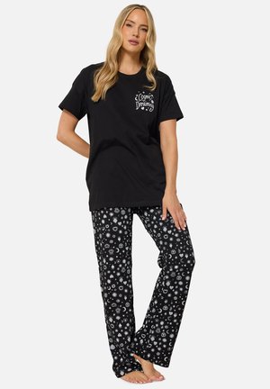 Mujer que lleva una camiseta negra "Cosmic Dreaming" y pantalones de pijama negros con patrones blancos de sol, luna, corazón y estrellas, de pie descalza.