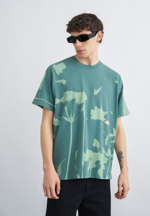 Jonge man met krullend haar en een zonnebril, gekleed in een groen tie-dye t-shirt en zwarte broek, staand tegen een effen lichte achtergrond.