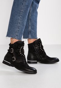 Black ankle boots made of leather with buckle straps, decorative studs, and a side zipper. The sole features a silver accent.