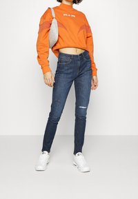 Orange kortärmad sweatshirt med logotyp, tillsammans med blå skinny jeans med ett hål och vita sneakers. En grå axelväska ger extra detalj.