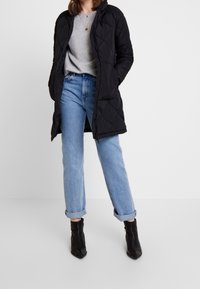 Černý prošívaný dlouhý kabát s kapucí, se zipovým uzávěrem, nošený přes šedý svetr a světle modré džíny s manžetami, doplněný o černé kotníkové boty.