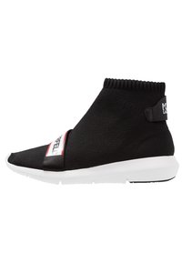 Black knit ankle-high sneaker with elastic cuff, featuring a white sole, a thin strap across the front with red and white accents, and logo detail.