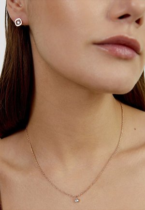 Primo piano del collo e della mascella di una donna che indossa una delicata collana d'oro con un piccolo ciondolo di diamante e orecchini a bottone con diamante abbinati.