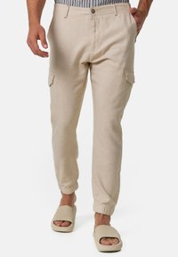 Beige Leinen-Cargohosen mit elastischen Bündchen, ausgestattet mit Seitentaschen und einem Knopfverschluss. Kombiniert mit neutralen Slip-On-Sandalen.