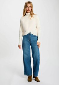 Maglione bianco a collo alto a coste con design incrociato abbinato a jeans blu a vita alta e gamba larga, e stivali alla caviglia marroni.