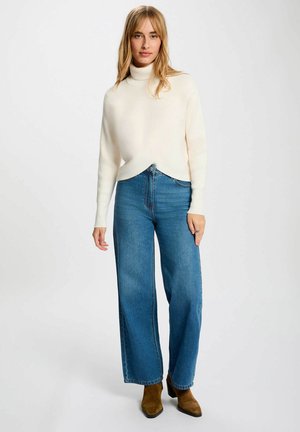 Maglione bianco a collo alto a coste con design incrociato abbinato a jeans blu a vita alta e gamba larga, e stivali alla caviglia marroni.