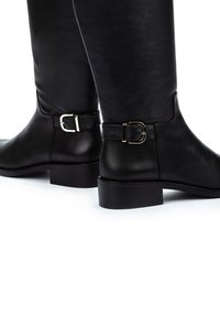 Botas de cuero negro hasta la rodilla con punta redondeada, tacón plano y detalle de hebilla plateada en el tobillo. Textura suave y diseño elegante.