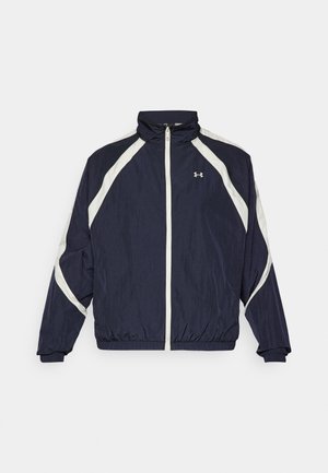 Navy tuulenpitävä takki, jossa on valkoiset vinoraidat, korkea kaulus, täysi vetoketjukiinnitys, joustavat hihansuut ja helmassa. Under Armour -logo rinnassa.