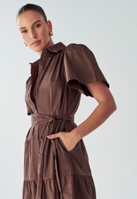 Rjava bombažna obleka s kratkimi napihnjenimi rokavi, sprednjim zapiranjem na gumbe, pasom in nabrano krilo. Ima stransko žepo. Gladka tekstura.