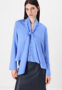 Bluză de culoare albastru deschis cu dungi verticale albe, cu legare frontală, închidere cu butoane și mâneci lungi. Țesătura pare netedă și ușoară.