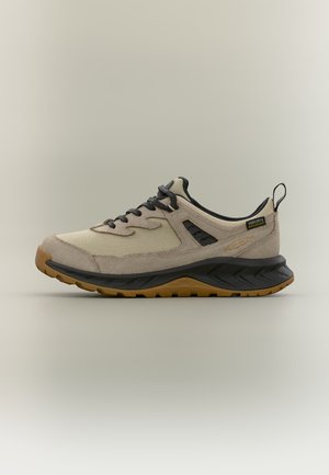 Μπεζ και μαύρο αδιάβροχο low-top sneaker με μαύρα κορδόνια, υφασμένο πέλμα και θηλιά στο πίσω μέρος, εμφανίζεται σε προφίλ πλευράς.