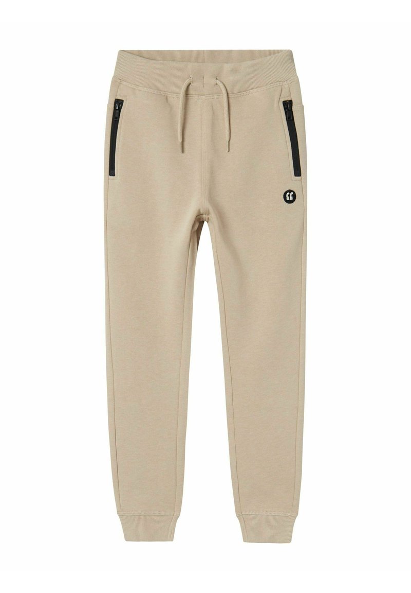 Beige Jogginghose aus weichem Material, mit schwarzen seitlichen Reißverschlusstaschen, einem gerippten Bund und einem kleinen Logo am linken Oberschenkel.
