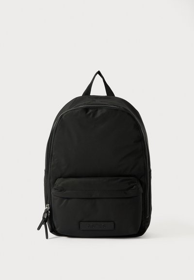 Sac à dos en nylon noir de forme arrondie, avec des compartiments zippés, une poche avant et une poignée supérieure. Présente un branding minimal à l'avant.