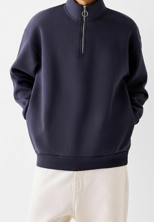 Person iført en mørkeblå sweatshirt med kvart-lynlås med ringtræk og hvide bukser, hænder i lommerne.