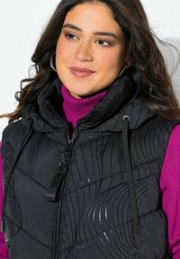 Mladá žena s tmavými vlnitými vlasy, která nosí stříbrné kruhové náušnice, fialový rolák a černou prošívanou vestu s vlnkovými vzory.